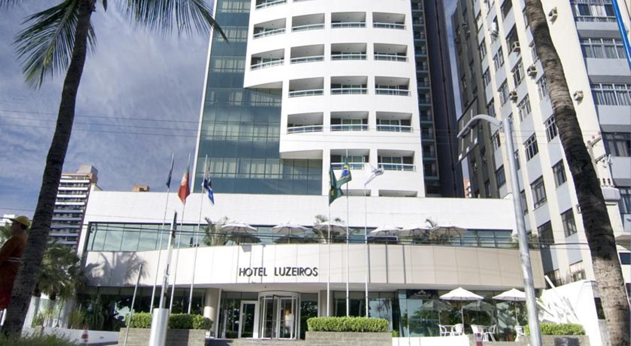 Hotel Luzeiros Fortaleza Exterior photo The photo shows the facade of a modern hotel named "Hotel Luzieros." The building features a sleek design with a combination of glass and concrete elements. Several flags are displayed in front, suggesting an international appeal. The entrance area i