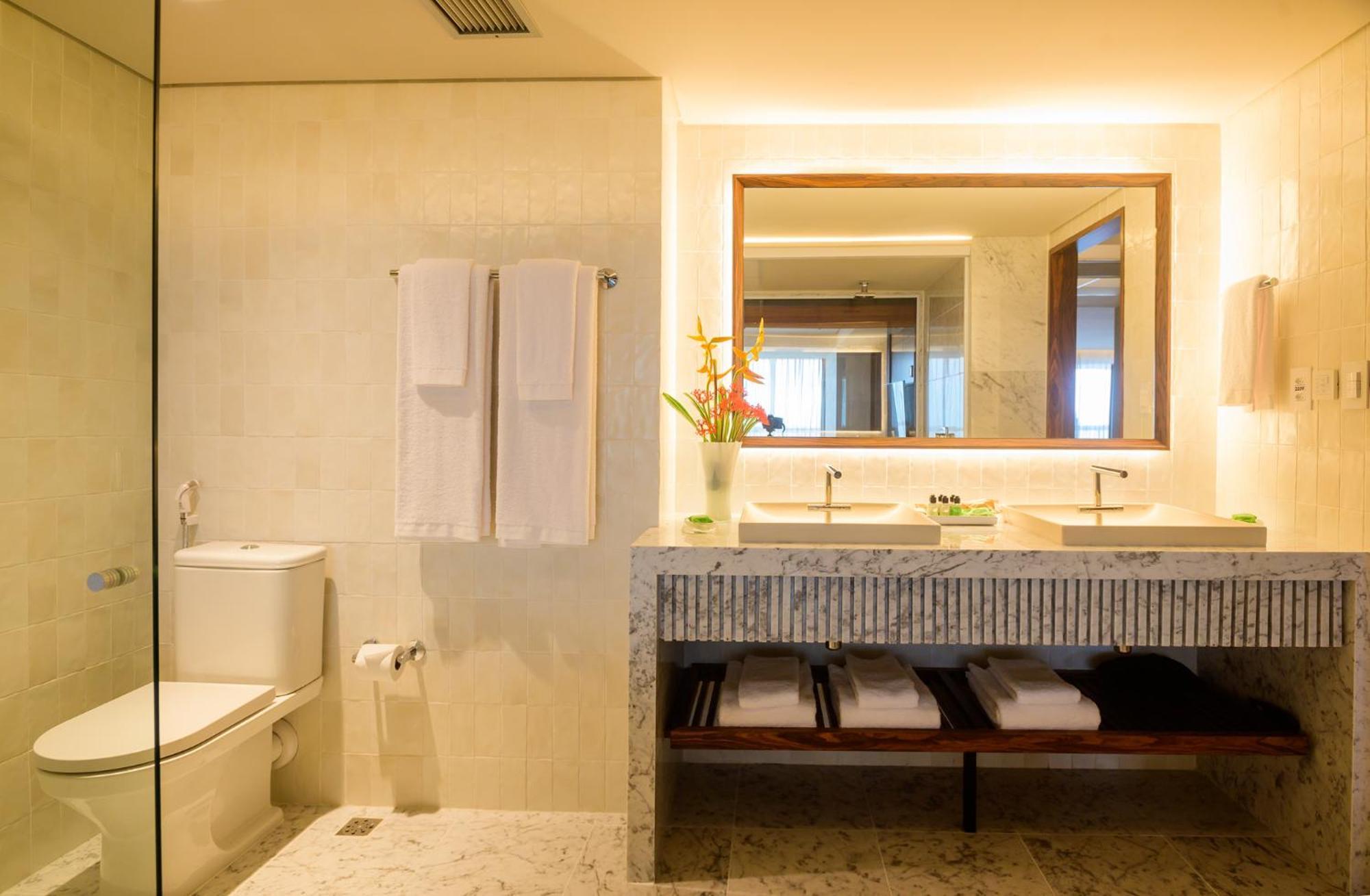 Hotel Luzeiros Fortaleza Exterior photo The photo shows a modern bathroom. It features a glass shower enclosure, a white toilet, and a double sink vanity made of stone. Above the sink, there's a large mirror with warm lighting around it. Hand towels hang neatly on a rack, and there are som